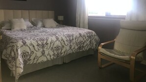 Un dormitorio con cama, una silla, mesita de noche, lámpara y ventana con cortinas.