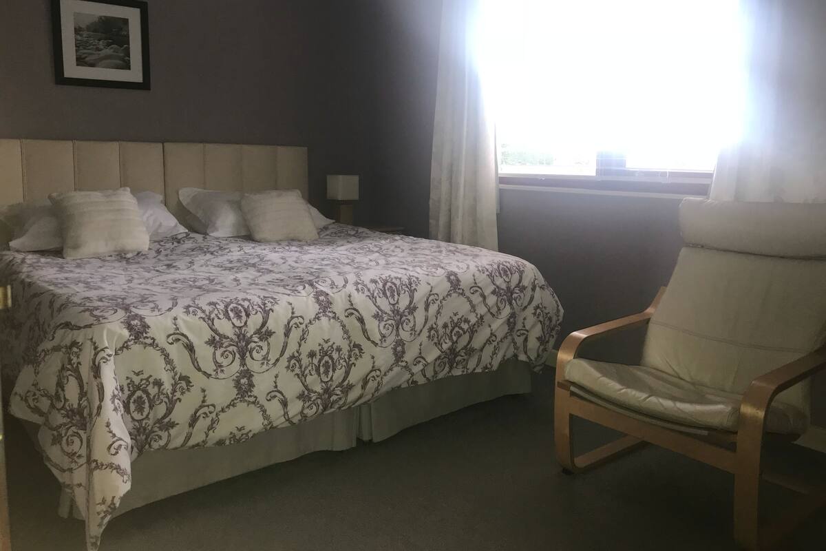 A bedroom with a bed, a chair, a nightstand, a lamp, and a window with curtains.