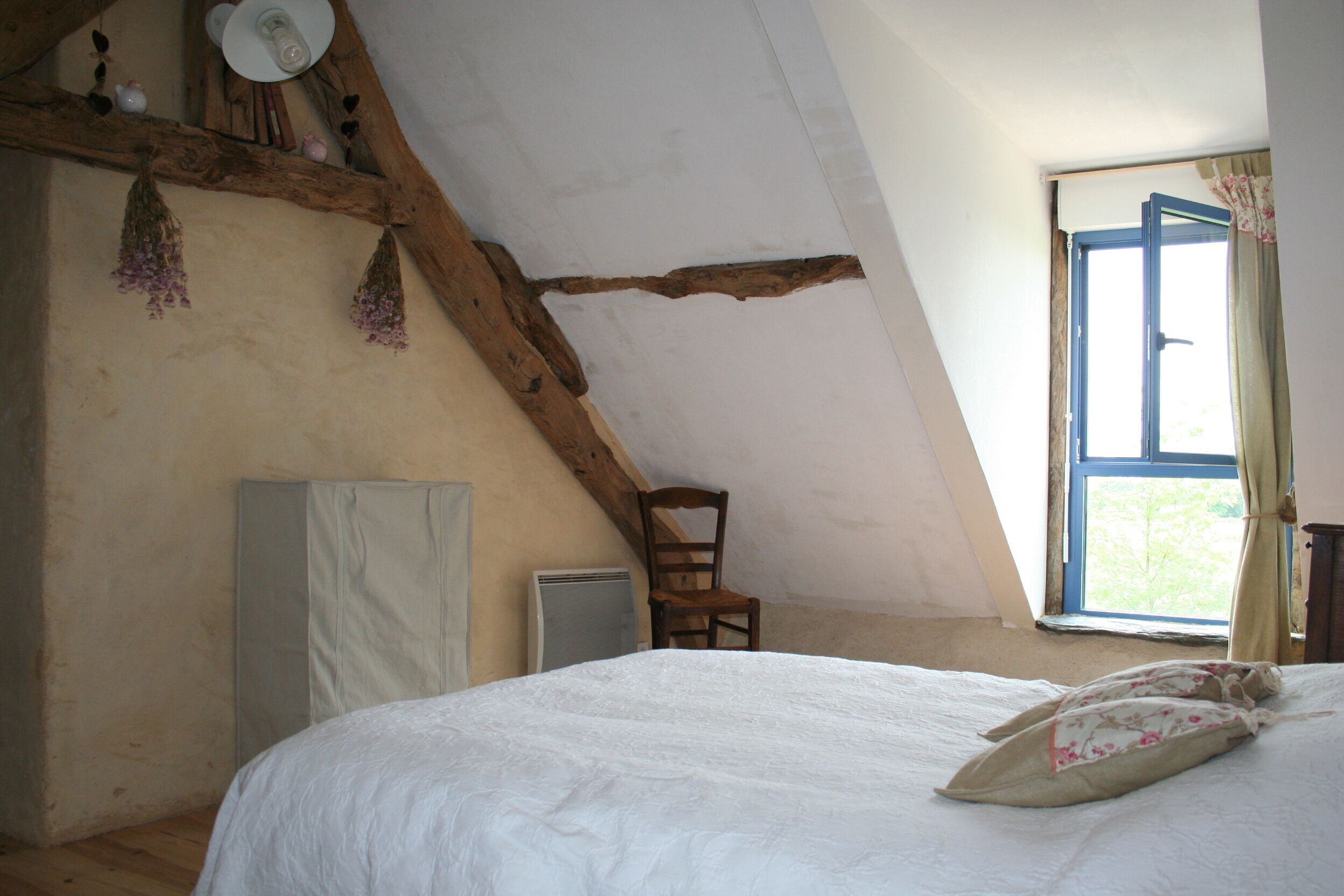 A bedroom with a sloped ceiling, a wooden chair, a bed with a white bedspread, and a window with curtains.