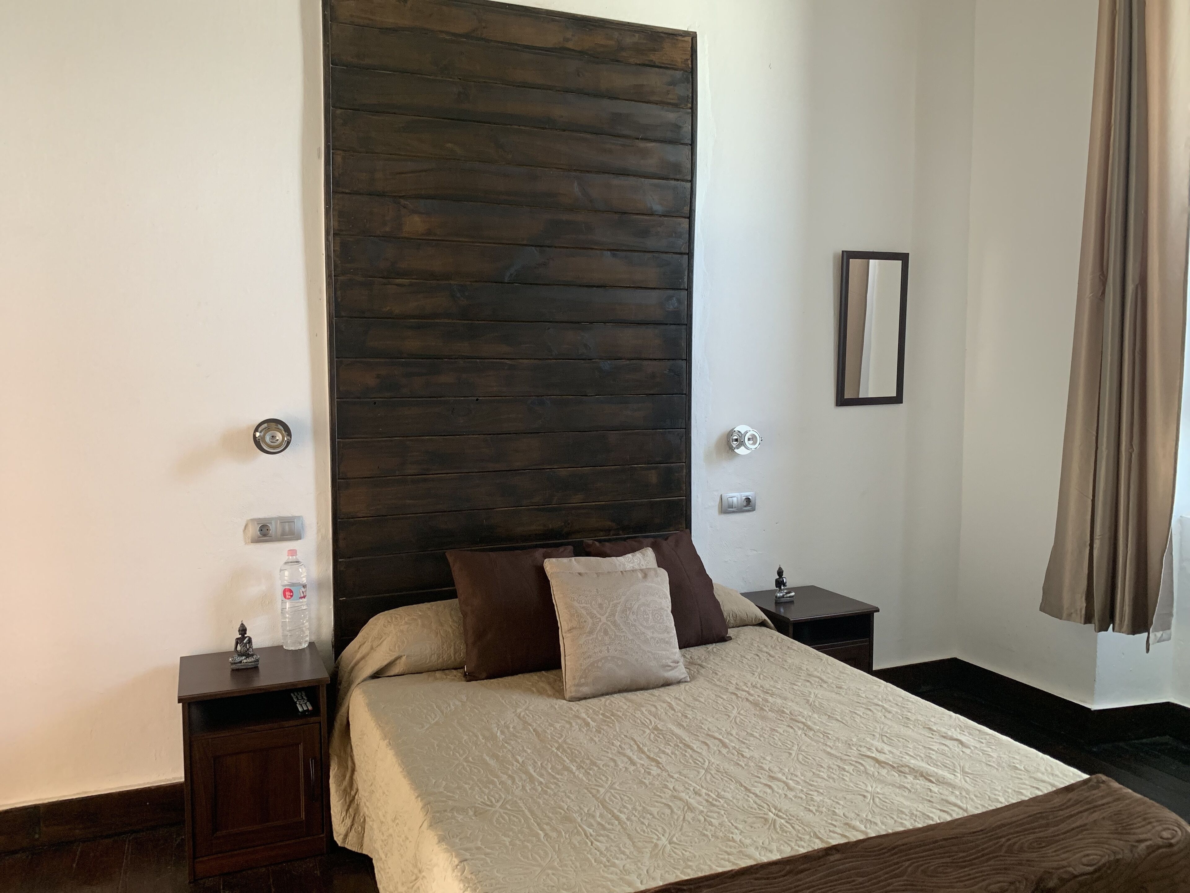 A bedroom with a wooden headboard, a bed with a beige bedspread, two bedside tables, and a nightstand with a bottle and a small statue.