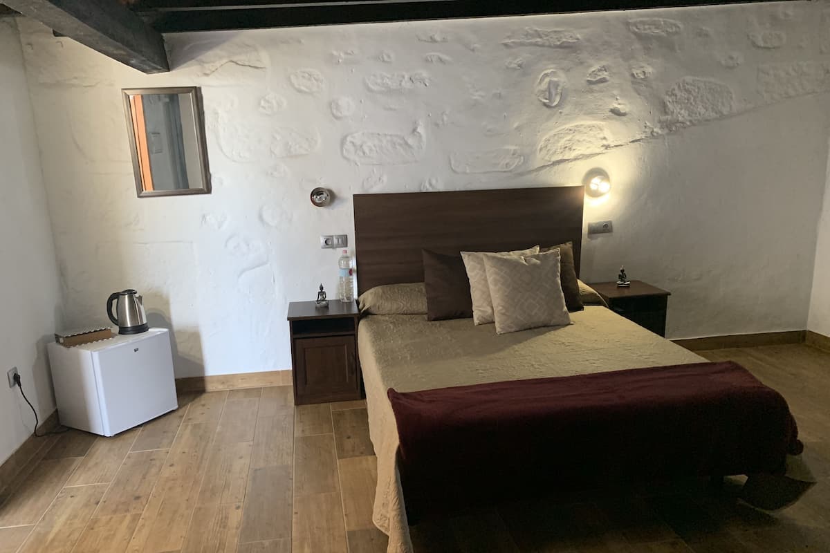 A bedroom with a wooden headboard, a bed with a red blanket, a nightstand with a lamp, a small white appliance, and a textured wall.