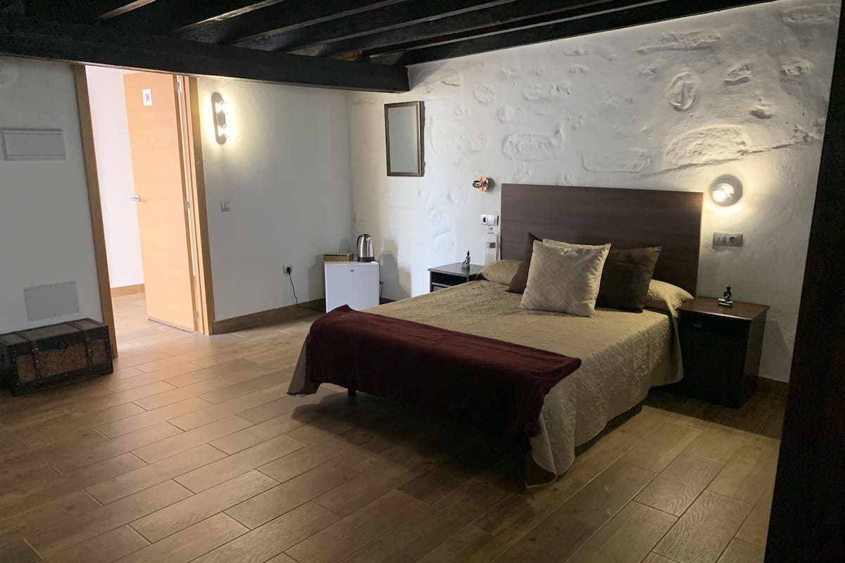 A bedroom with a wooden floor, a bed with a red blanket, a nightstand with a lamp, and a wall-mounted shelf.