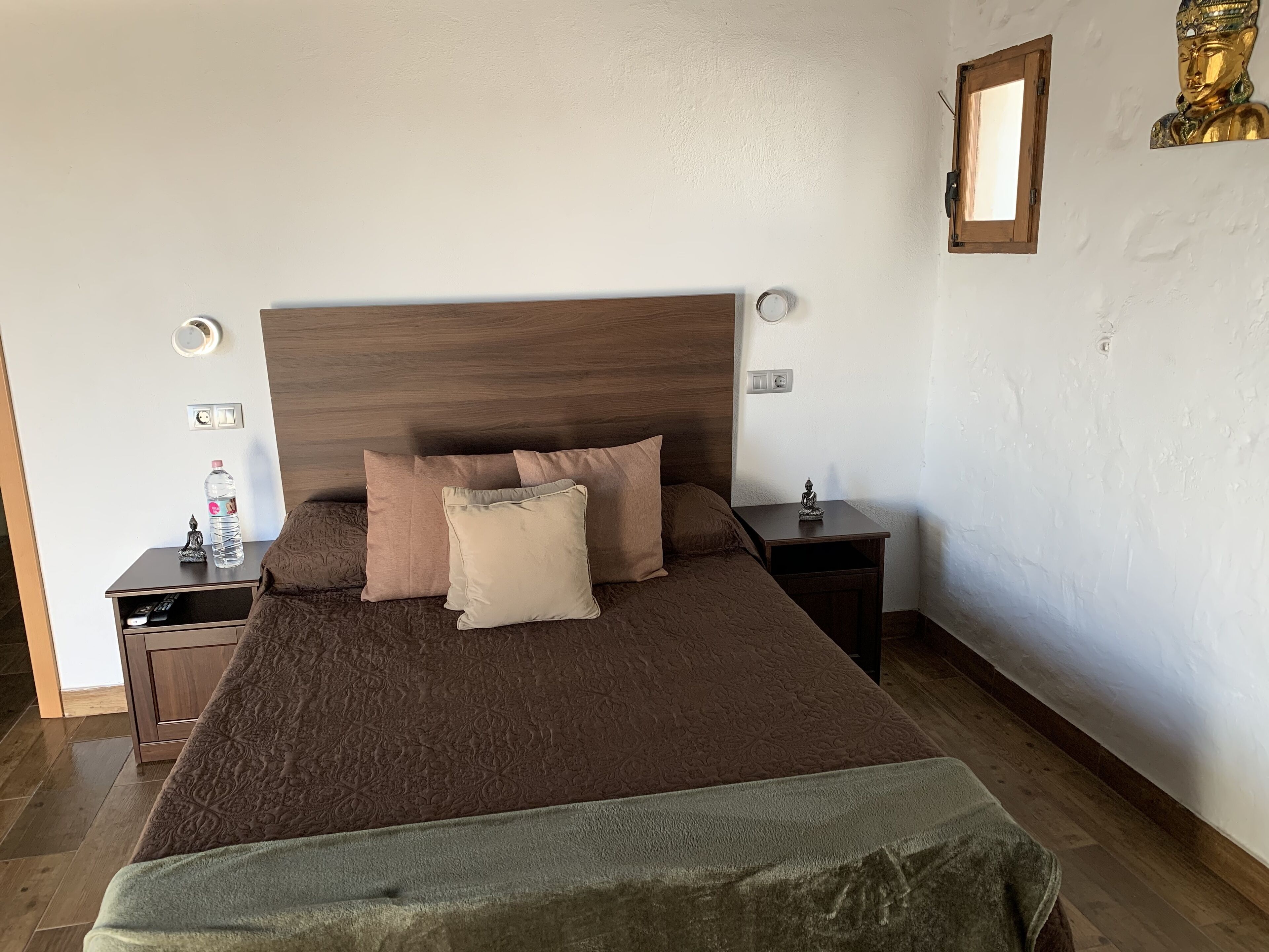 A neatly made bed with a brown headboard, a bedside table with a water bottle and a small statue, and a wall-mounted Buddha statue.