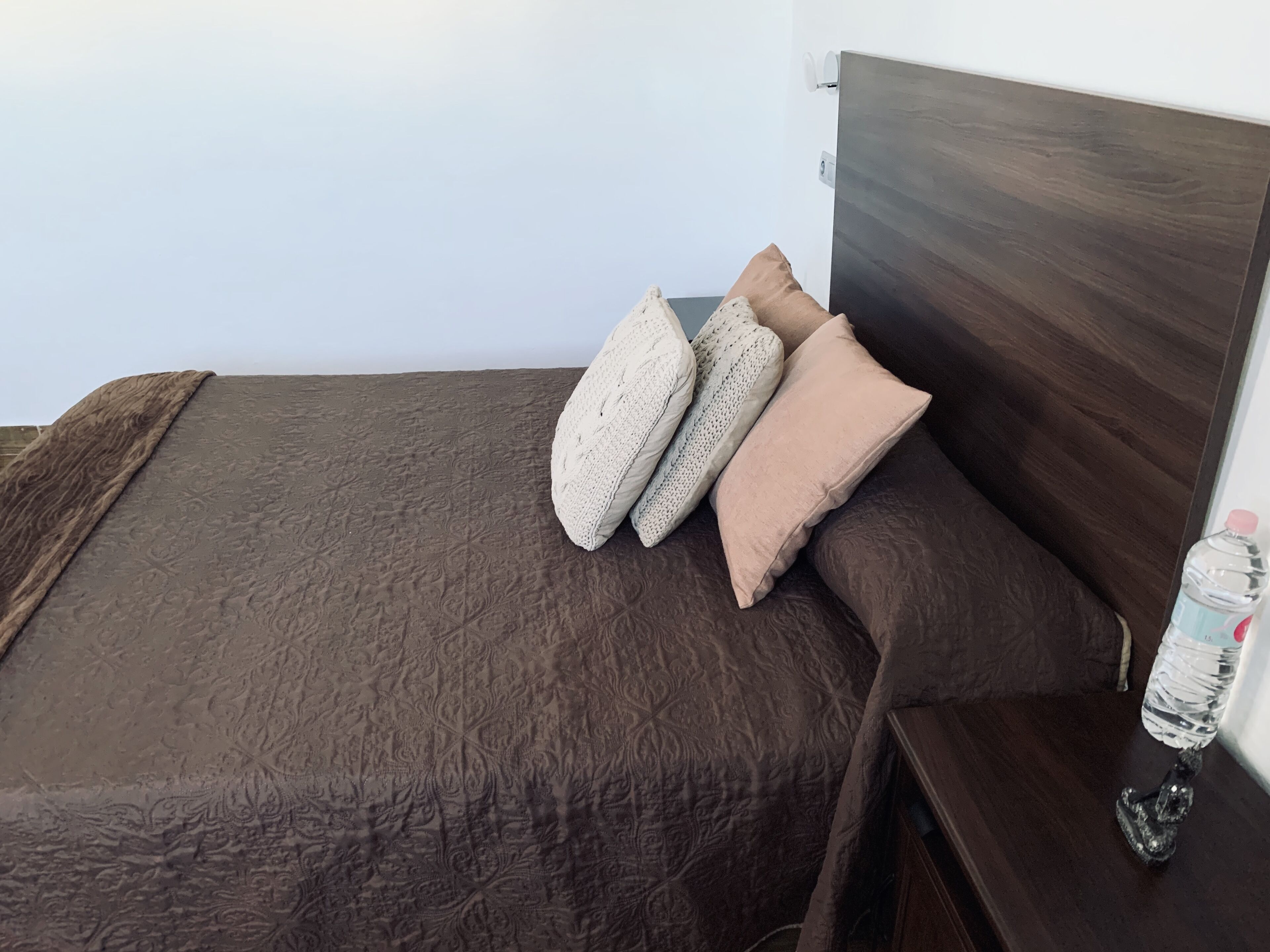 Un lit recouvert d’une couverture marron à texture, agrémenté de deux coussins à motifs, et une bouteille d’eau posée sur une table de chevet en bois.