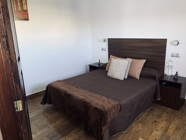 A neatly made bed with a brown bedspread and two pillows, a wooden headboard, a nightstand with a water bottle and a Buddha statue, and a wooden floor. - Valtisya House Pool & Airport (Telde)