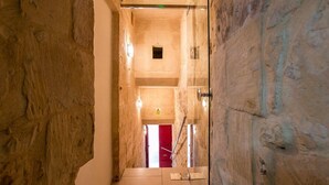 A narrow stone corridor with a red door at the end, a green emergency exit sign, and a warning sign on the wall. - Vallettastay Banoffee (Valletta)