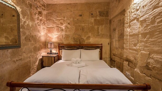 A stone-walled room with a wooden bed, a nightstand with a lamp, and a mirror on the left wall. - Vallettastay Banoffee (Valletta)