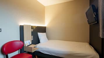 A small, neatly arranged bedroom with a single bed, a red chair, a bedside table with a lamp, and a television mounted on the wall.