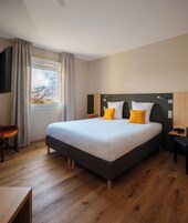 Una habitación de hotel moderna con una cama grande, suelo de madera, un escritorio y una ventana con cortinas.