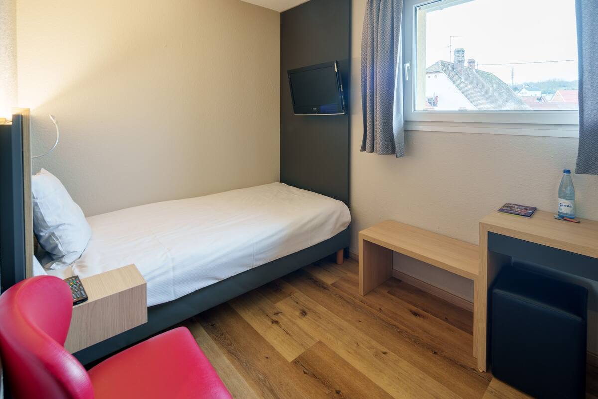 A compact hotel room with a single bed, a small desk, a chair, and a TV mounted on the wall.