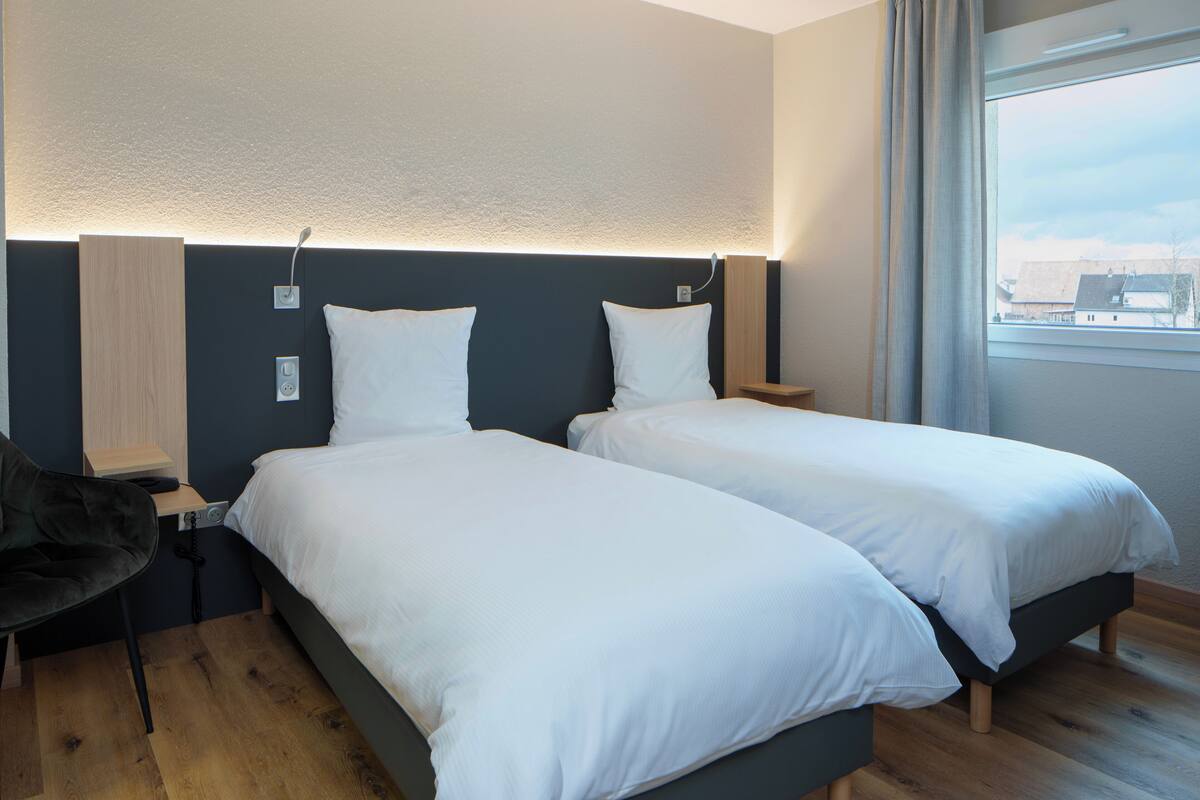 A hotel room with two single beds, a desk, a chair, and a large window with a view of buildings.