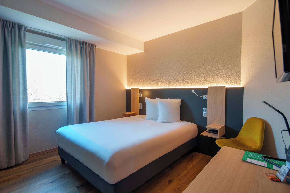 A hotel room with a large bed, a desk, a chair, a nightstand, and a wall-mounted lamp.
