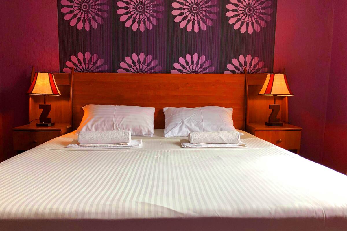 Un lit bien fait, avec deux oreillers et deux serviettes pliées, flanqué de deux tables de chevet avec des lampes, le tout adossé à un mur orné d’un motif floral.