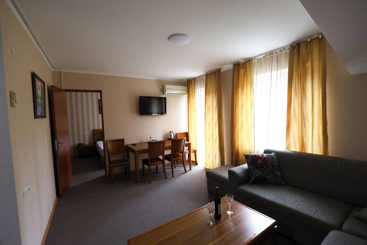 Un salon comprenant un canapé, une table à manger avec des chaises, un téléviseur fixé au mur et une fenêtre avec des rideaux.