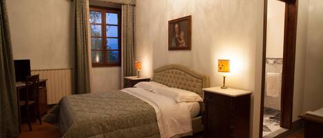 Un dormitorio con cama, mesita de noche, escritorio, silla, ventana con cortinas, un cuadro en la pared y la puerta del baño.