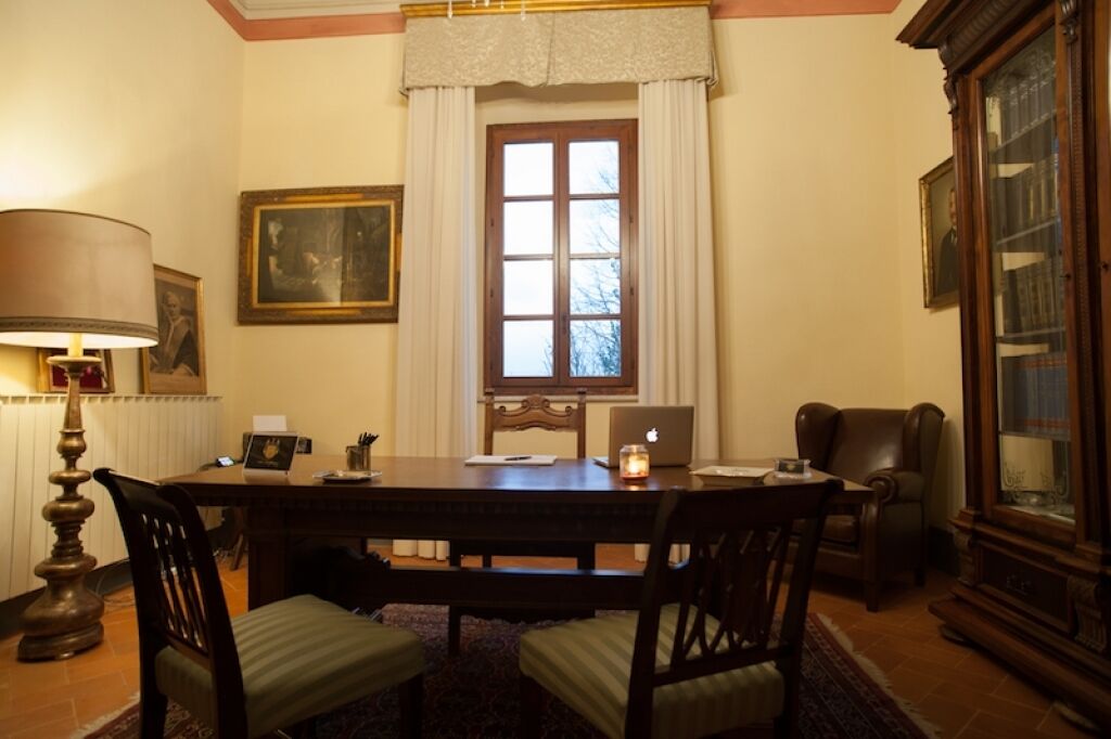 A dining area with a wooden table, chairs, a lamp, a window with curtains, and a bookshelf.