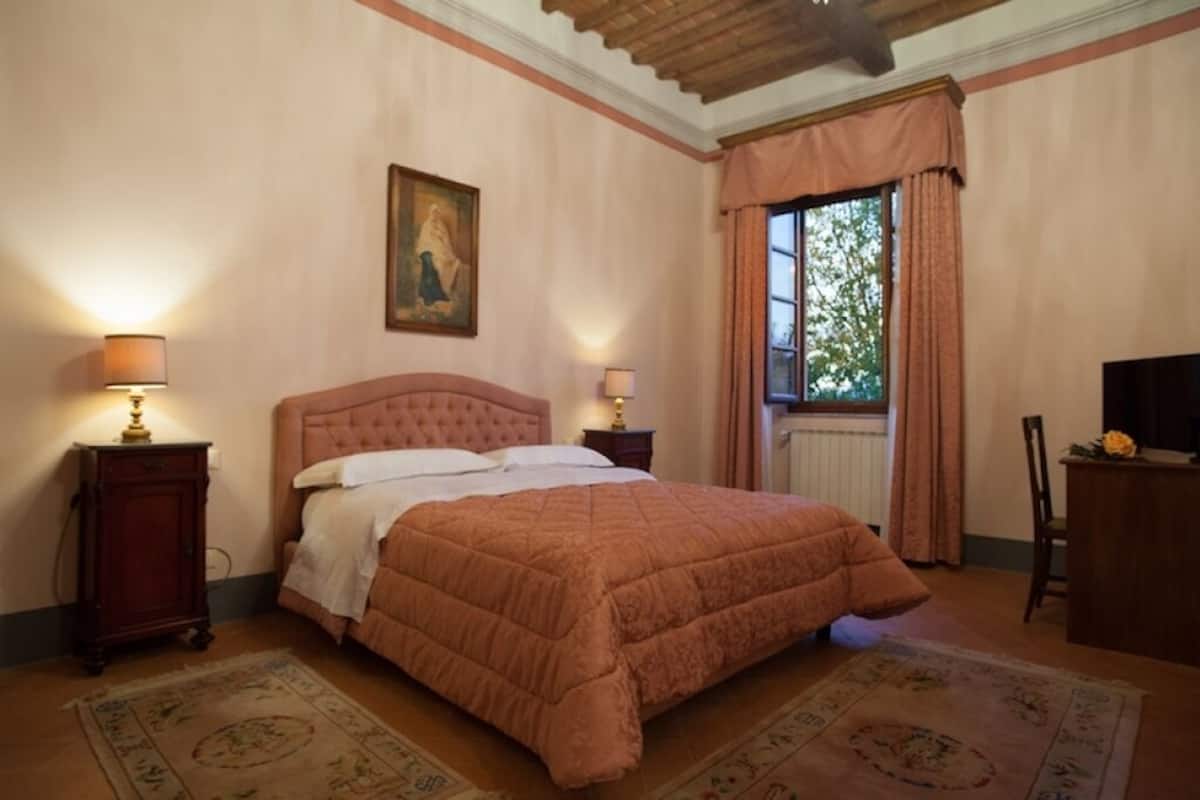 Een slaapkamer met een groot bed, twee nachtkastjes met lampen, een schilderij aan de muur, een raam met gordijnen en een bureau met stoel.