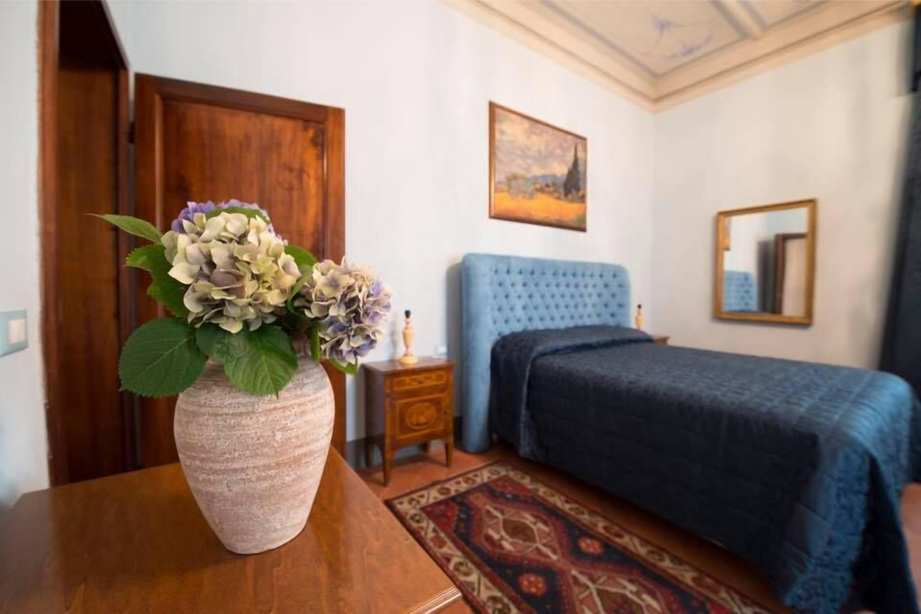 A bedroom with a blue upholstered bed, a wooden nightstand with a lamp, a vase with flowers, and a patterned rug.