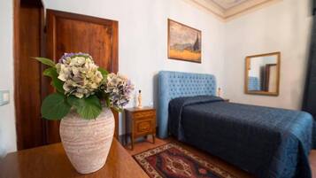 A bedroom with a blue upholstered bed, a wooden nightstand with a lamp, a vase with flowers, and a patterned rug.
