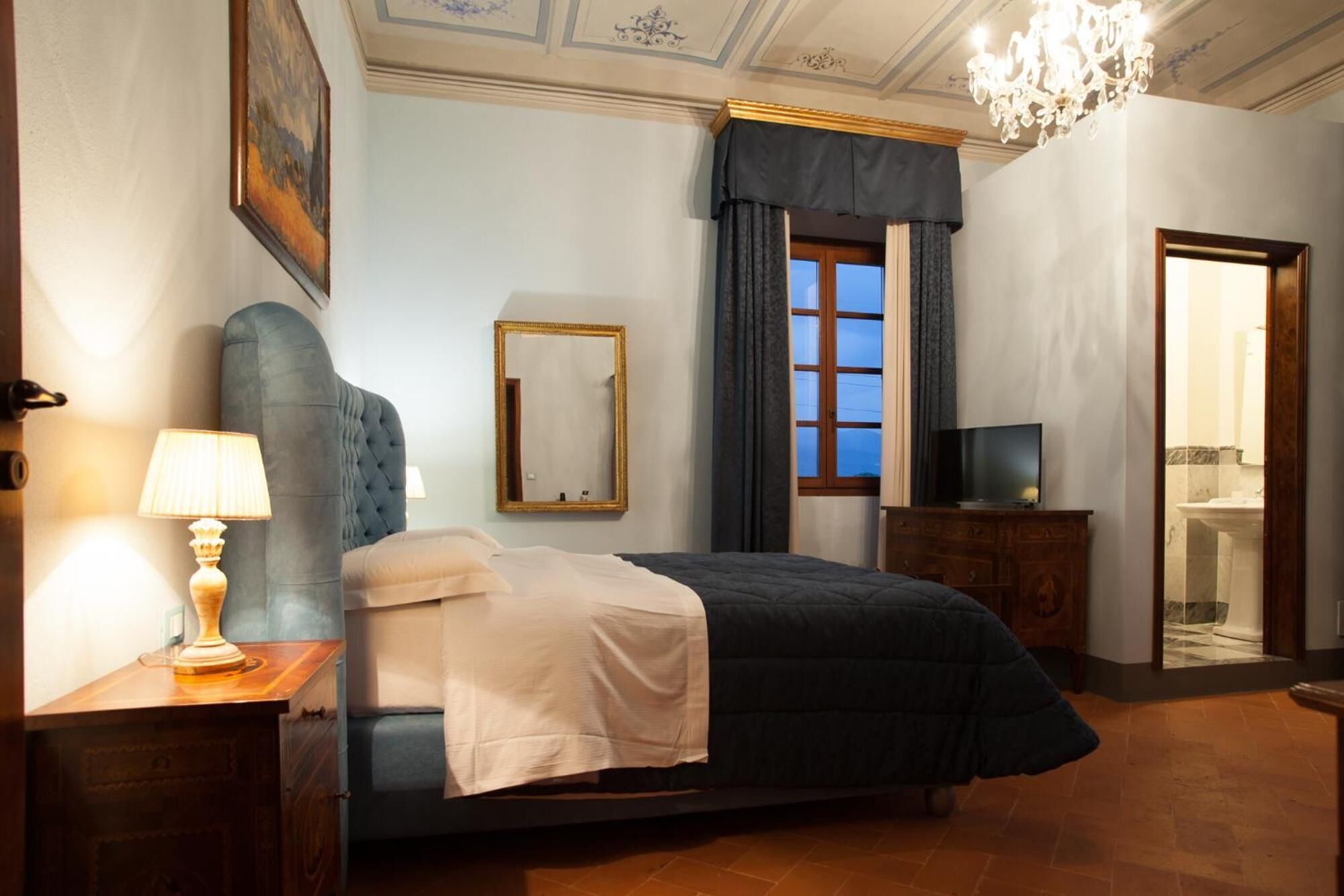 A bedroom with a large bed, a bedside table with a lamp, a mirror, a TV, and a chandelier.