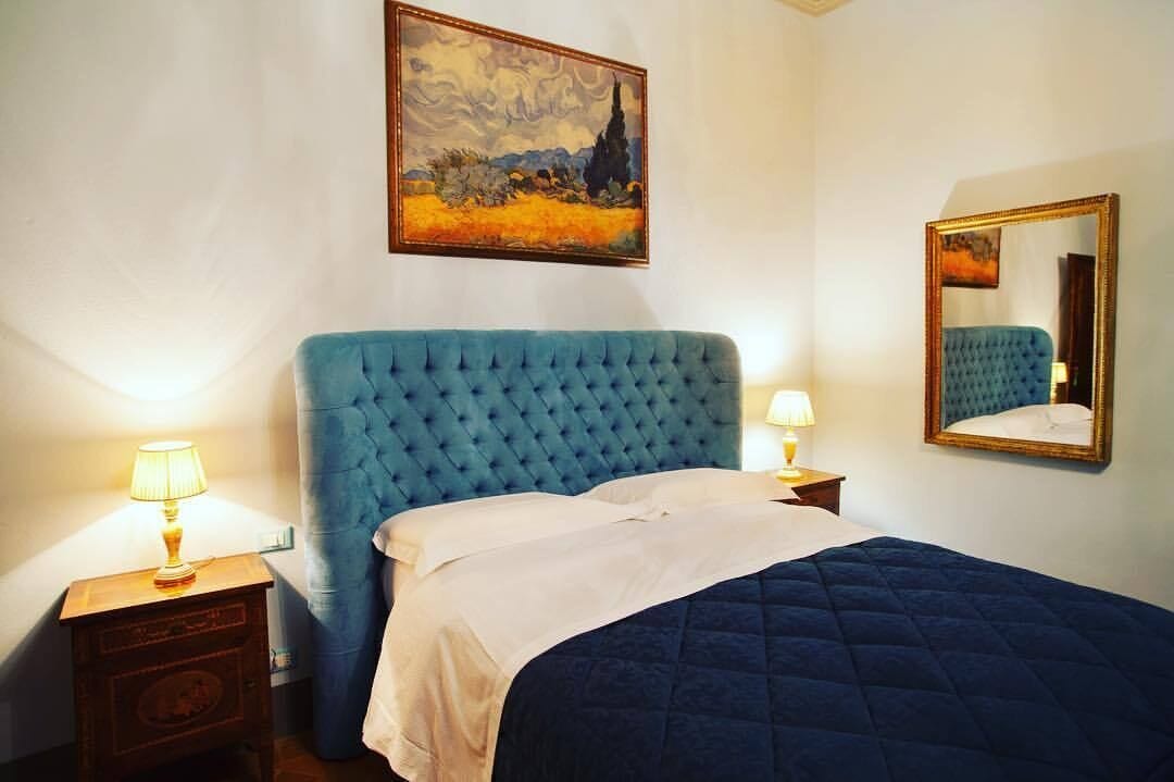 Una camera da letto con una grande testiera imbottita blu, una coperta bianca, due abat-jour, un comodino in legno, un quadro incorniciato sopra il letto e uno specchio appeso alla parete.
