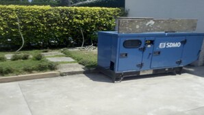 Una máquina industrial azul con la etiqueta 'SDMO' en un patio con seto y sendero adoquinado.