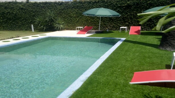 Una piscina rectangular con borde blanco, rodeada de césped artificial verde. Hay dos sillas reclinables rojas y un paraguas verde.