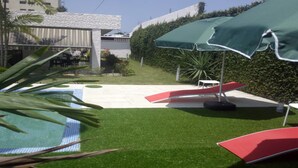 Zona de piscina con césped artificial, un tobogán rojo y una sombrilla verde.
