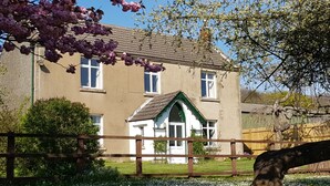 Une maison à deux étages avec une façade à pignon, une clôture en bois au premier plan et un jardin où poussent différentes sortes de fleurs.