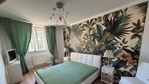 Um quarto com colcha verde, cabeceira branca, mesa de cabeceira com luminária, TV fixada na parede e papel de parede tropical.
