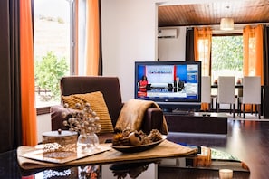 Una sala de estar moderna con un televisor mostrando un noticiero deportivo, un rincón de estar cómodo y vistas al exterior.