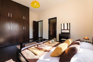 Un dormitorio moderno con una cama grande, un armario de madera oscura, un espejo y una mesita de noche con una lámpara.