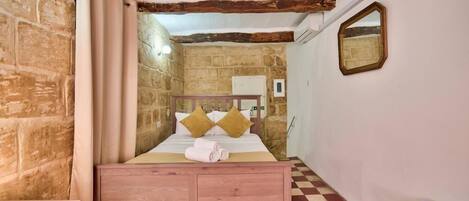 Een slaapkamer met een geruite vloer, een houten bed met kussens, een houten nachtkastje, een spiegel en een stenen muur.
