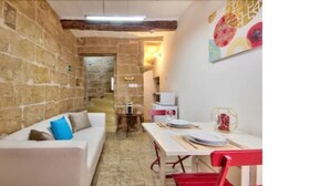 Un pasillo estrecho con pared de piedra, un sofá blanco con un cojín azul, una mesita con una silla roja y un microondas sobre una repisa.
