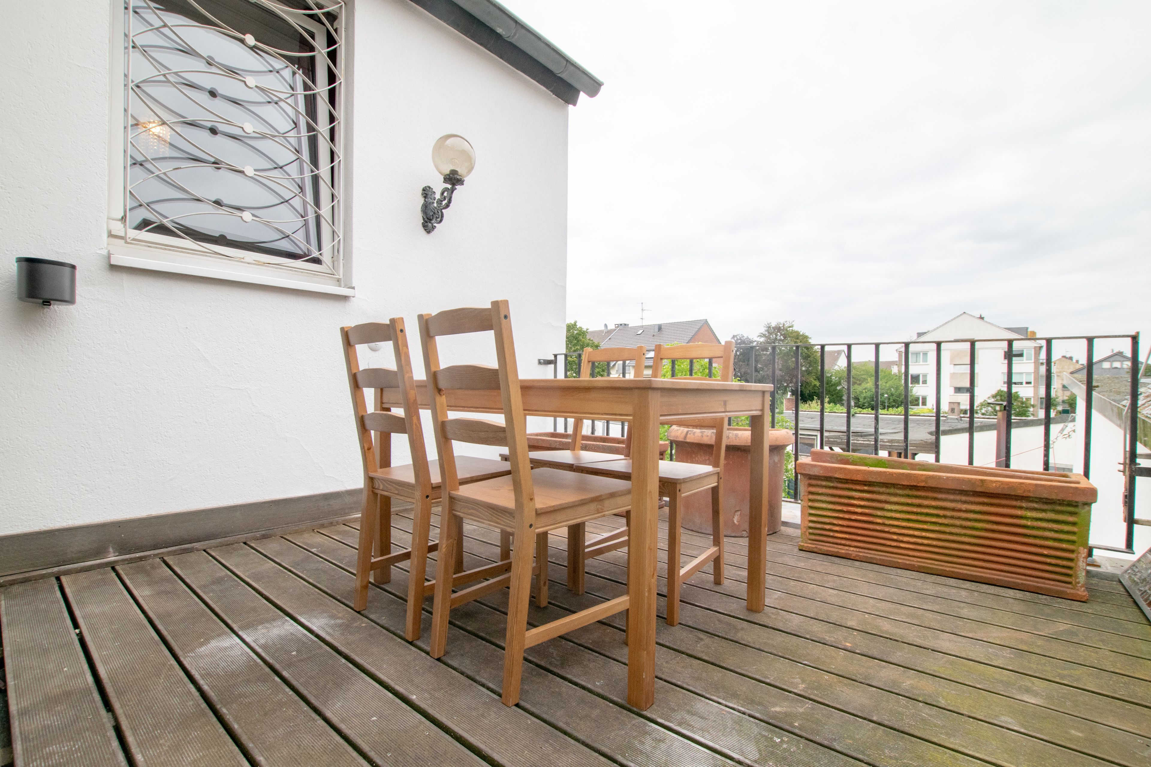 Una terraza de madera con una mesa, sillas, una maceta y un ventanal con un celosía decorativa.