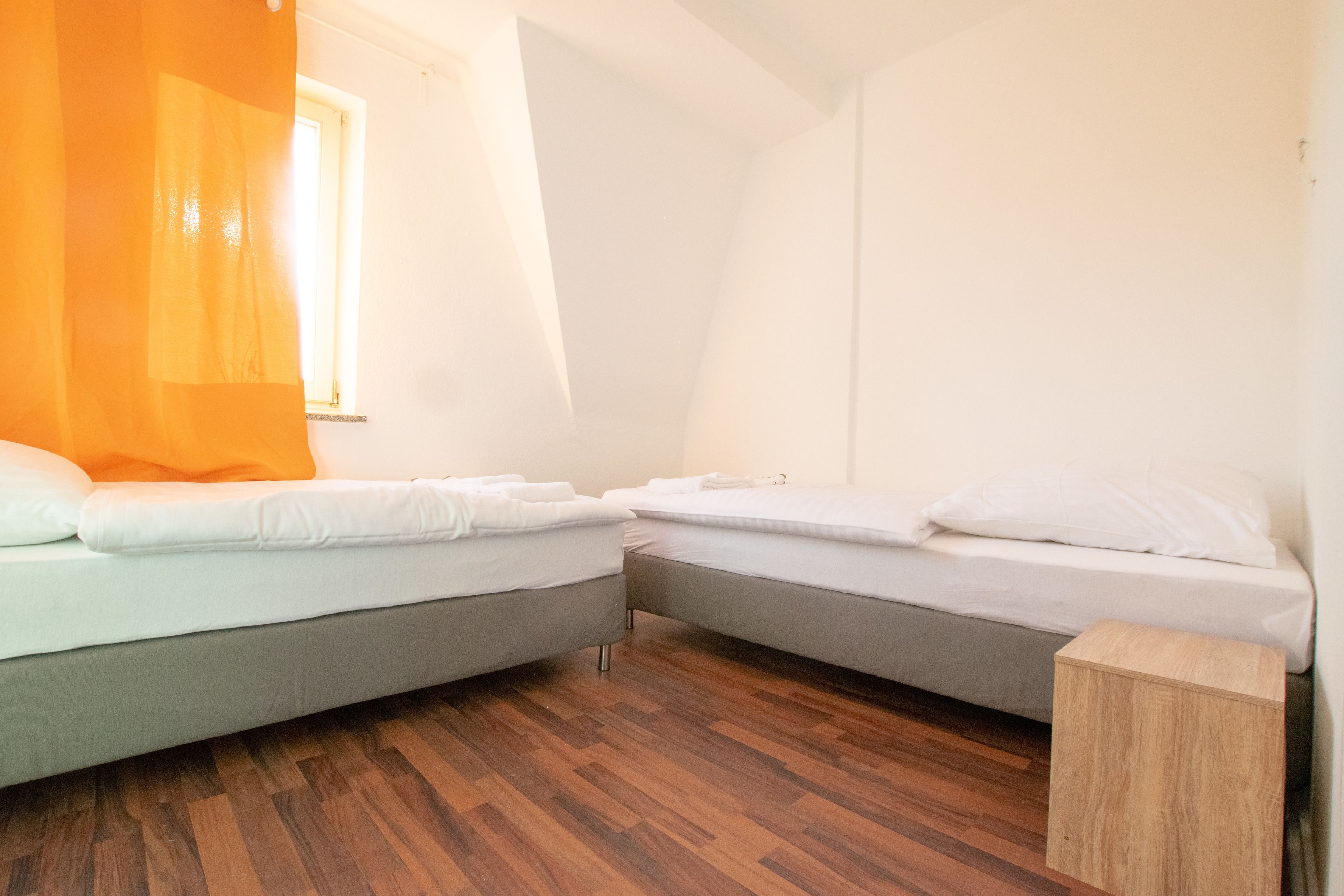 A room with two beds, a wooden floor, and a window with a curtain.