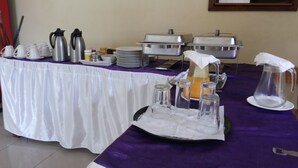 Montaje de buffet con diferentes recipientes de comida, vasos y una jarra sobre una mesa cubierta con mantel blanco.
