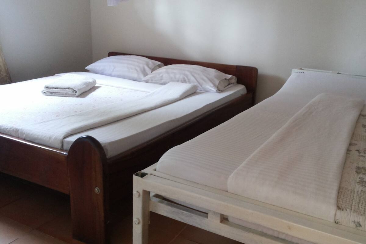 Habitación con dos camas, una más grande y otra más pequeña, ambas con ropa de cama y almohadas blancas.