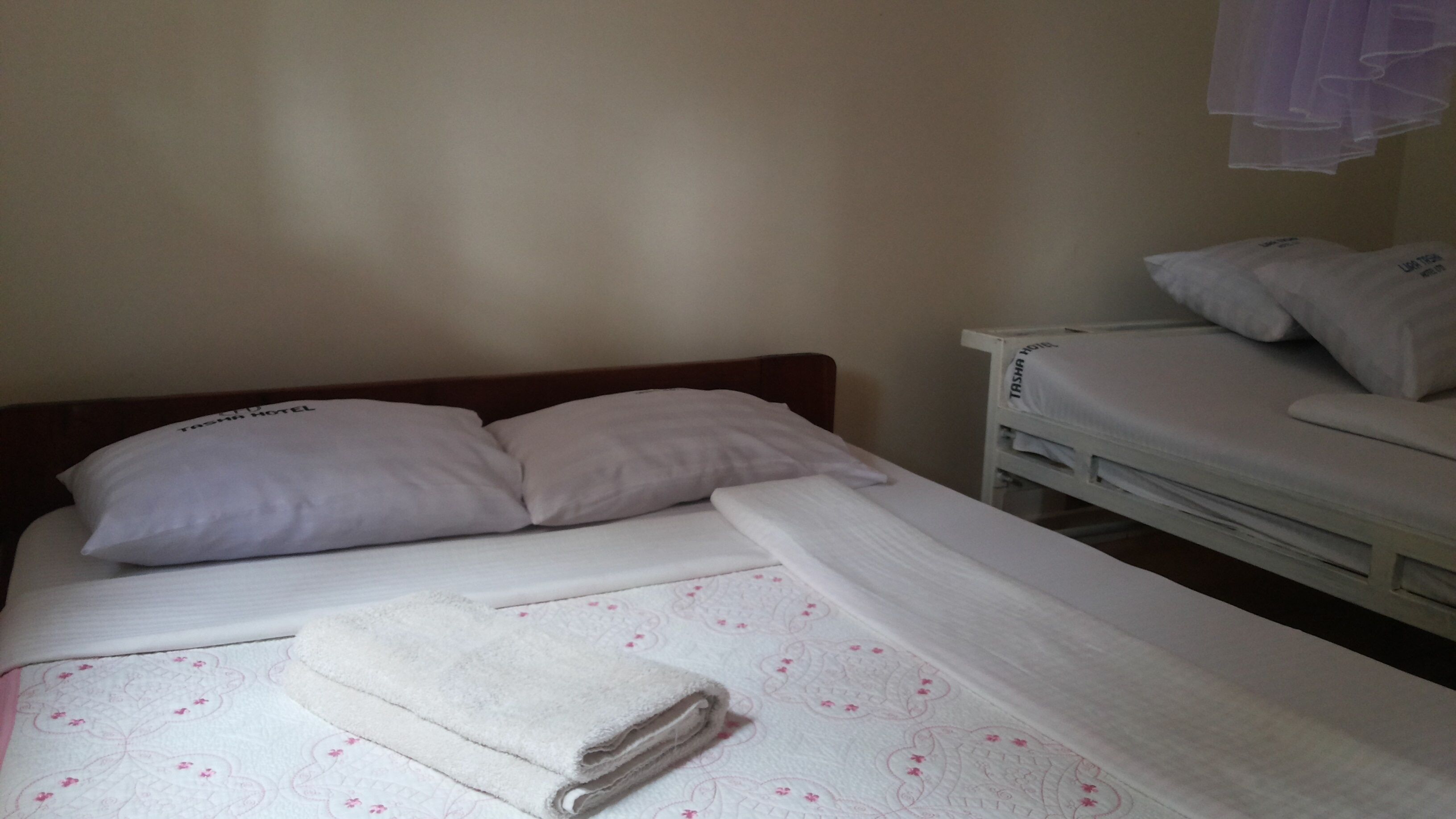 Una cama individual con cabecera de madera, dos almohadas, una toalla blanca doblada con cuidado y un colchón blanco con estampado floral.