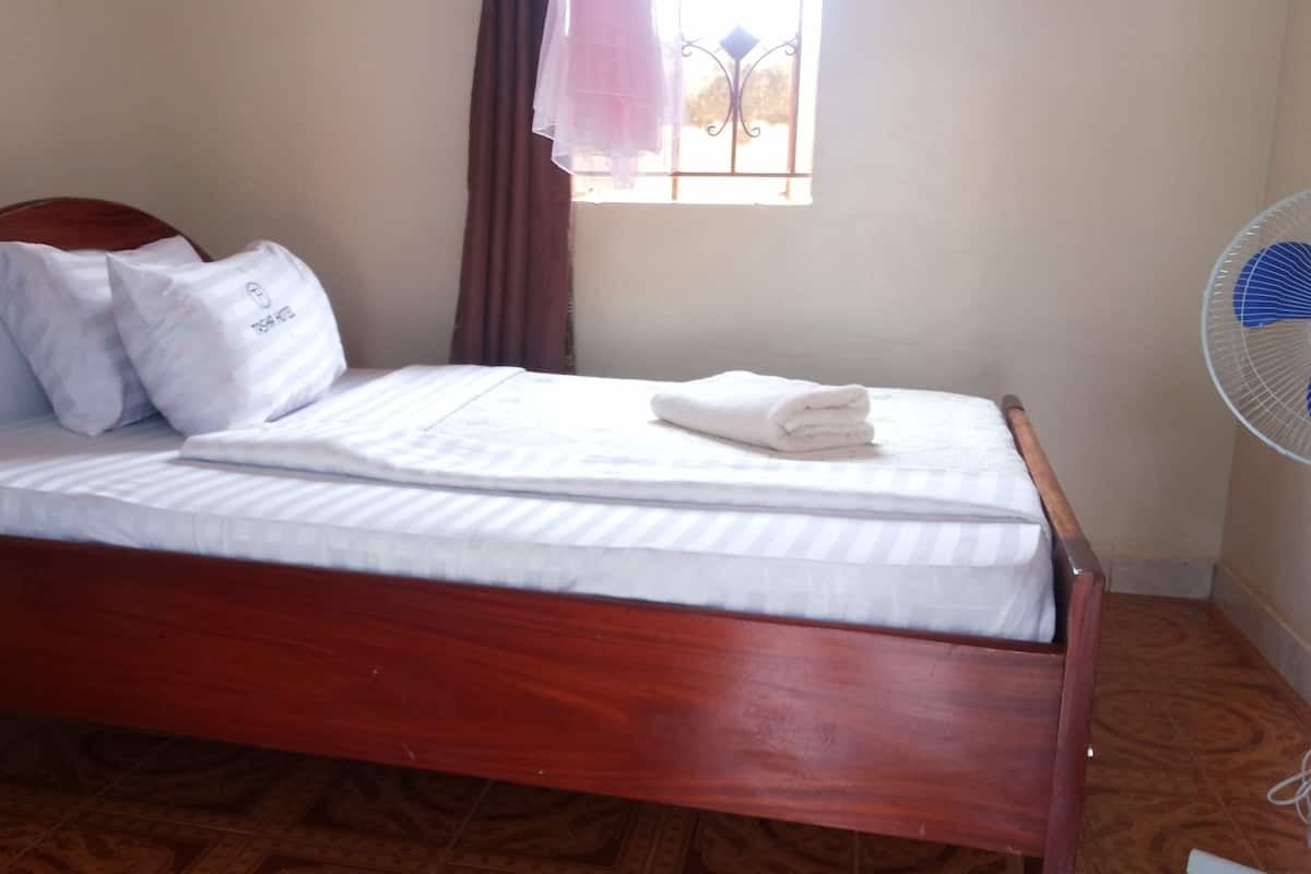 Una cama de madera con ropa de cama blanca, una ventana con cortinas y un ventilador de pie.