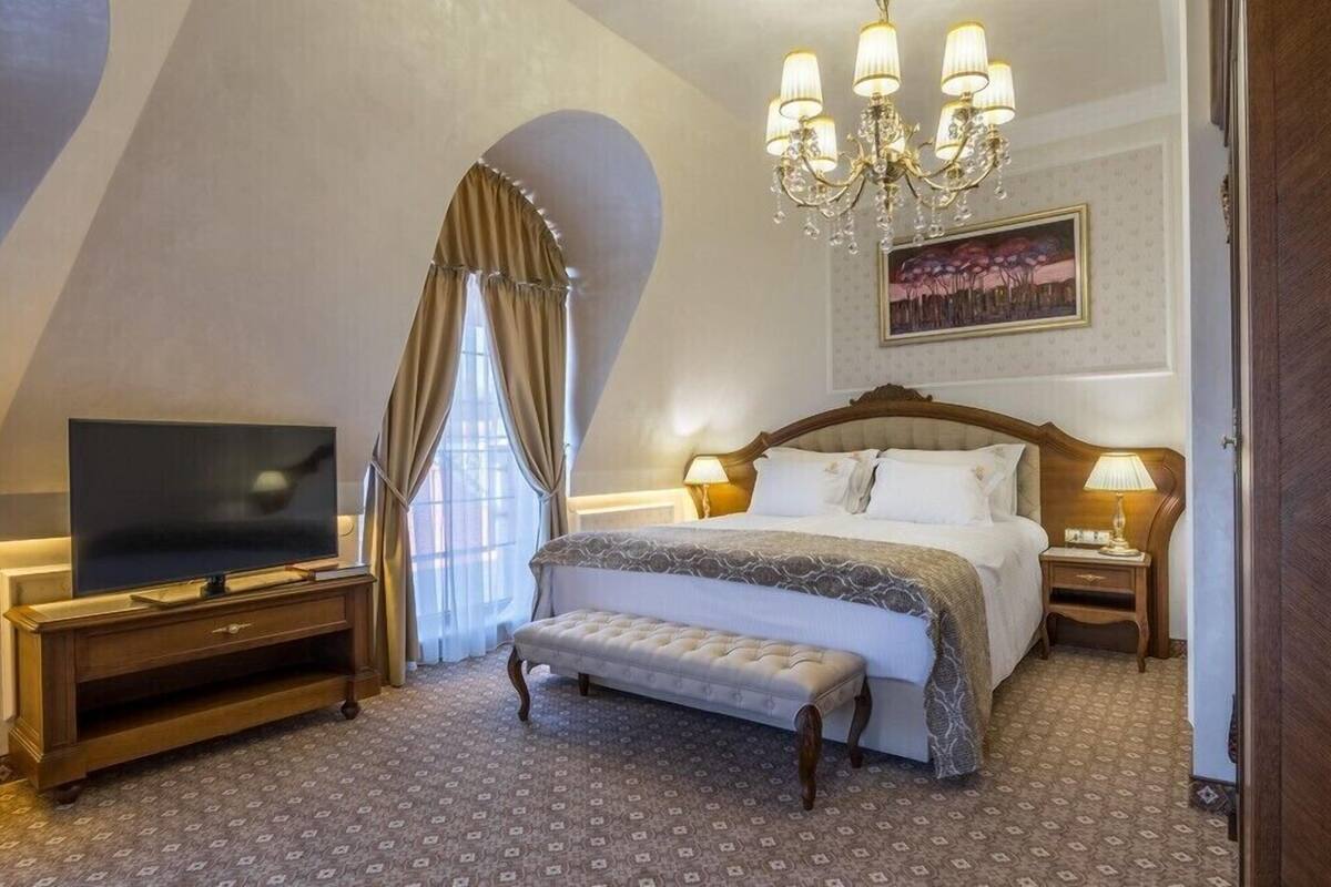 A bedroom with a large bed, a television on a wooden stand, a bench, and a chandelier.