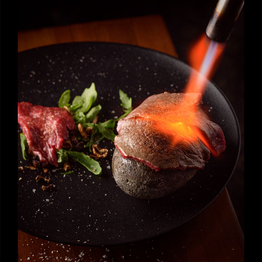 Un bistec a la parrilla sobre una losa de piedra con un plato negro, rodeado de vegetales y especias.