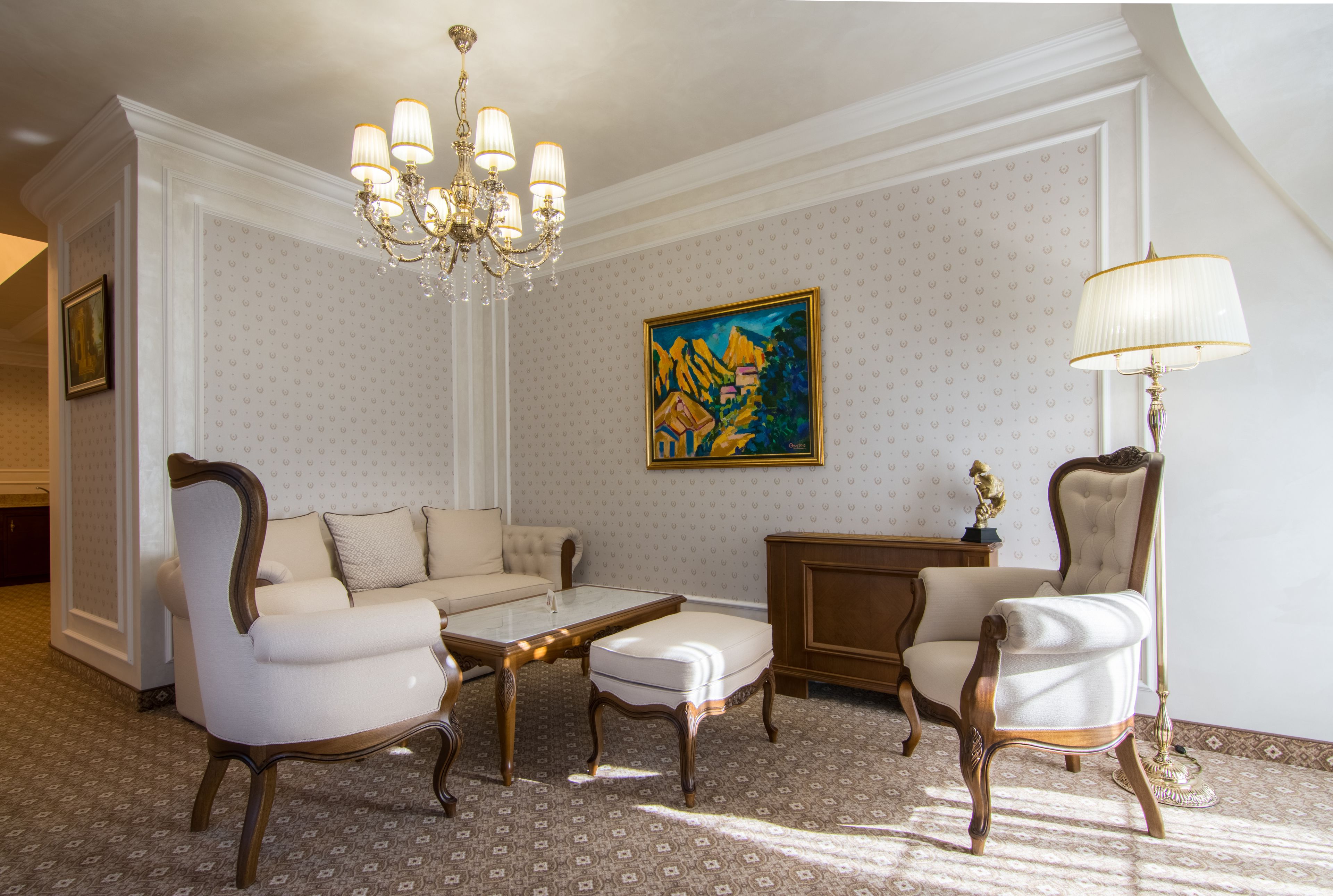 A room with a white patterned wallpaper, a chandelier, a painting, a sofa, a chair, and a lamp.