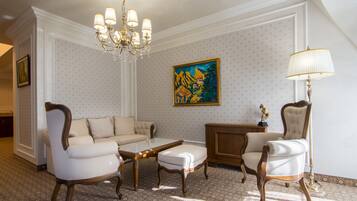 A room with a white patterned wallpaper, a chandelier, a painting, a sofa, a chair, and a lamp.