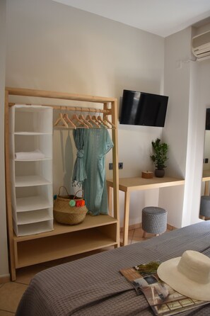 Et soveværelse med en seng, et fjernsyn ophængt på væggen, en træskab med plads til ophængning, et lille bord med en plante og et natbord med et magasin og en hat.