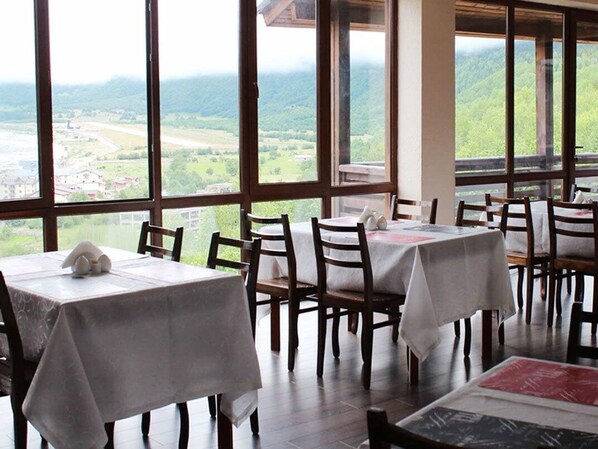 A restaurant with tables set for dining, large windows offering a view of a valley, and wooden chairs. - Hotel Banguriani (Mestia)