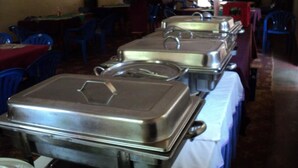 Un buffet con diverse stoviglie di acciaio inossidabile disposte su tavoli, sedie di plastica blu e un pavimento rivestito di moquette.