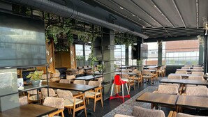 Un restaurant moderne avec des tables et des chaises en bois, de grandes fenêtres et des plantes d’intérieur.