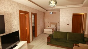 A living room with a flat-screen TV, a green sofa, a wooden cabinet, and a bed with a white headboard. - Van Hotel (Van)