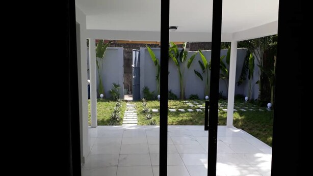 Un patio moderno con suelo de baldosas, un sendero que lleva a una reja, y un jardín con diversas plantas y una pequeña fuente.
